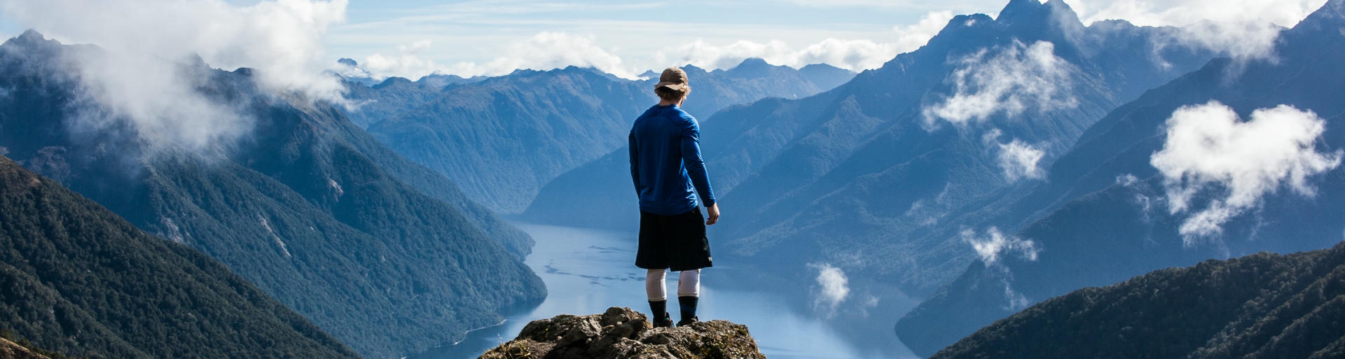 Mann steht auf Felsvorsprung in Neuseeland und schautin die Ferne auf einen Fjord.
