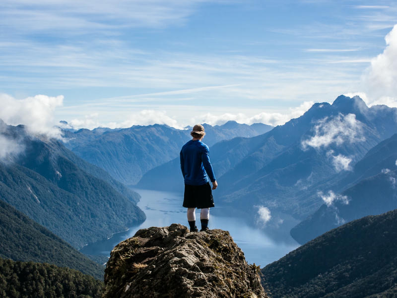 Mann steht auf Felsvorsprung in Neuseeland und schautin die Ferne auf einen Fjord.